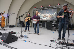 Outdoor stage with four musicians playing bass, guitar, banjo, and violin; a Tonewood backdrop looms behind them.