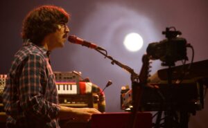 Side view of a male singer in a plaid shirt singing into a red microphone while playing a keyboard on a dimly lit stage, with studio gear in the background.