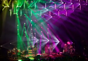 Live band on stage under vibrant green, pink, and purple lighting with multiple musicians and instruments visible.