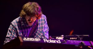 Musician wearing a plaid shirt and glasses playing a Roland JUNO-60 keyboard on stage, illuminated by purple lighting.