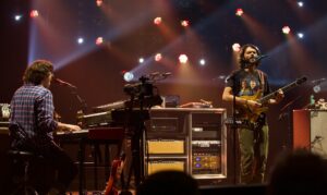 Two musicians on stage: a keyboard player in a plaid shirt on the left and a guitarist singer on the right under colorful stage lights.