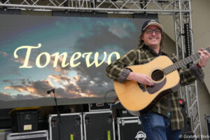Male guitarist in a plaid shirt and cap playing an acoustic guitar on a stage with a large screen backdrop showing cursive text behind him.