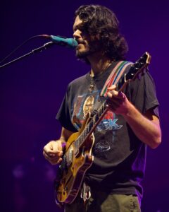Male guitarist performing on stage, singing into a teal microphone with a purple-lit background.