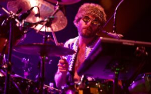 Drummer with a fur hat and round sunglasses performing on stage among cymbals and drums.
