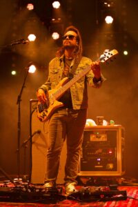 Bass guitarist wearing sunglasses and a denim jacket performs on a warmly lit stage with amplifiers behind him.