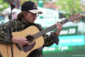 Young man playing an acoustic guitar outdoors, wearing a black cap and glasses.