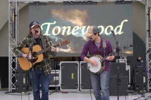 Two musicians perform on a stage: left guitarist sings into a mic, right guitarist plays a banjo; a large screen behind shows 'Tonewood'.