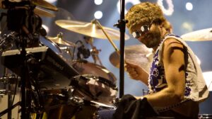 Drummer on stage wearing sunglasses and a white floral shirt, playing a drum kit with cymbals in view.
