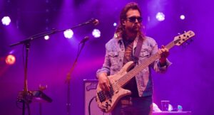 Bass guitarist wearing sunglasses and a denim jacket performs on a purple-lit stage with a light-colored bass guitar.