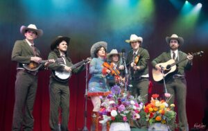 Group of six musicians in cowboy outfits performing on a stage with microphones and flowers in front.