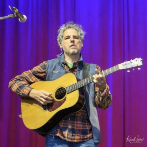 Male musician playing an acoustic guitar on stage, wearing a plaid shirt and denim vest, with a purple curtain backdrop.