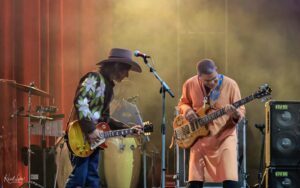 Two guitarists perform on a stage bathed in warm colored lights; one wears a wide-brim hat and floral shirt, the other in an orange outfit playing bass guitar.