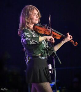 Female violinist on stage wearing a green embroidered jacket, playing a violin with a bow under purple stage lighting.