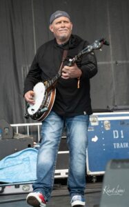 Male musician in a black hoodie and Yankees cap playing a banjo on stage with gear cases in the background.