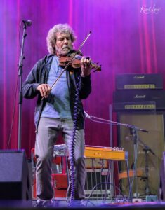 Older man with gray hair and beard playing a violin on a purple-lit stage, microphone in front and guitar amps in the background.