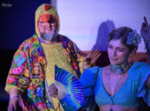 Two people dancing at a colorful party: a person in a bright chicken costume and a woman in a blue dress with a flower in her hair, holding a fan.