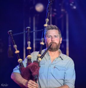 Bearded man in a light blue shirt on stage, speaking into a microphone, with a large maroon plush bear in the foreground and stage lights behind him