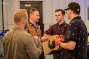 Three men converse at an indoor event, one holding an acoustic guitar and wearing a patterned shirt.