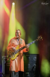 Male bassist in an orange robe playing a yellow bass on stage under colorful yellow and red lights, with gear in the background, smiling toward the audience.