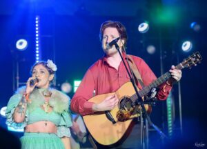 Male guitarist-vocalist and female singer perform on stage under blue concert lighting.