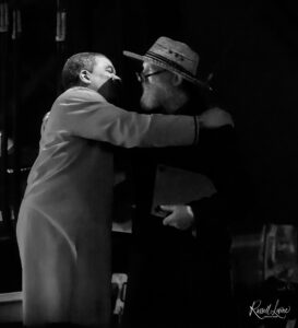 Two people embracing in a dark, black-and-white photo, one wearing a wide-brimmed hat visible on the right side.