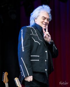 Musician with white hair smiling on stage, wearing a black embroidered jacket; two guitars visible at the bottom left.