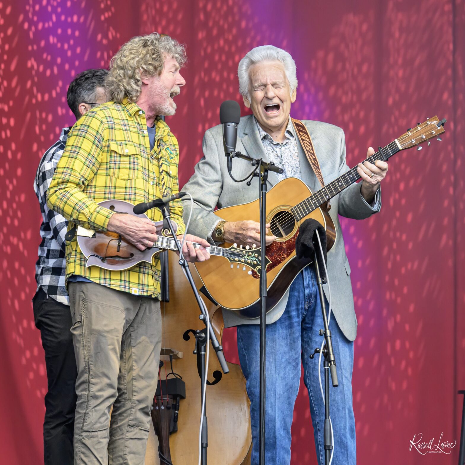 Three male musicians perform on a stage, two with guitars, singing into microphones against a red-purple backdrop? (live performance)