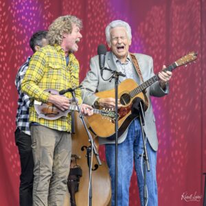 Three male musicians perform on a stage, two with guitars, singing into microphones against a red-purple backdrop? (live performance)