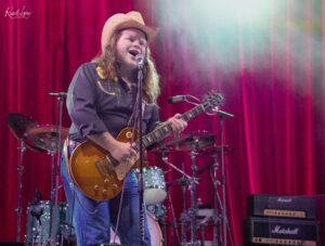 Male guitarist wearing a straw hat sings into a microphone on a red-velvet stage, playing a sunburst Les Paul guitar.