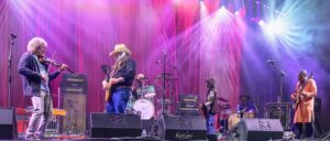 Band performs on a colorful lit stage with guitars, drums, and a violinist under pink and purple lights.