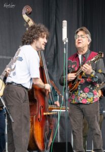 Two male musicians on stage: one with curly hair playing a double bass, the other in a floral shirt singing into a vintage microphone while playing a guitar, during a live performance.