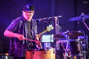 Drummer wearing glasses and a black cap plays drums on a purple-lit stage.
