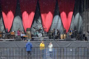 A live concert stage with red drapes, musicians and instruments set up, and audience barrier in front on a rainy day