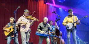 Five musicians perform on a stage with red and blue lights: guitarist on the left, violinist next to him, a player with a blue hammered dulcimer in the center, an upright bass player behind, and a banjo player on the right.