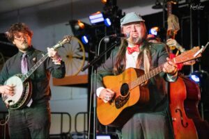 Musician singing into a microphone while playing an acoustic guitar on stage, with a banjo player beside him under stage lights.