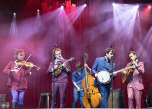 A group of musicians on a stage playing violins, guitars, and a bass under pink and purple stage lights with a red curtain backdrop.
