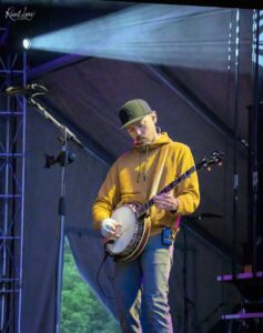 Musician in a mustard hoodie playing a banjo on a dimly lit outdoor stage with blue and purple lighting.