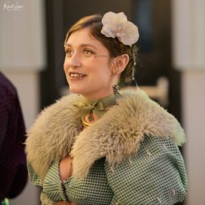 Young woman with a white flower in her hair, wearing a green gingham shirt and fluffy coat, smiling gently on her right side.