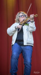 Violinist performing on stage in a white jacket and sunglasses against a red curtain backdrop.