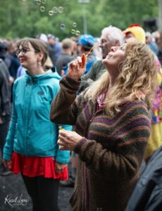 Woman with curly hair blowing bubbles using a wand at an outdoor crowd event, people in jackets in the background.