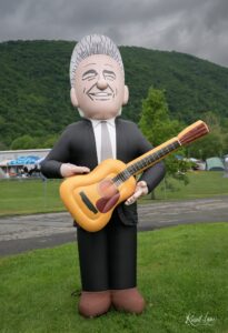 Large inflatable figure in a suit playing an acoustic guitar at an outdoor festival, with tents and green hills in the background.