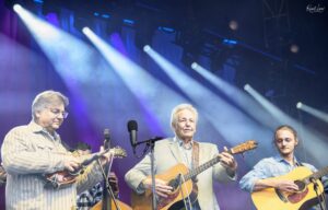 Four male musicians perform on a stage with acoustic guitars and bright blue-purple spotlights.