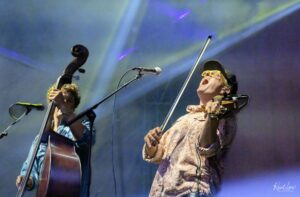 Two musicians on stage: a bassist left, a vocalist with a violin on the right, singing into a mic under colorful stage lights.