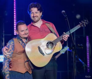 Two male musicians on stage with an acoustic guitar, smiling under purple stage lights with a microphone nearby.