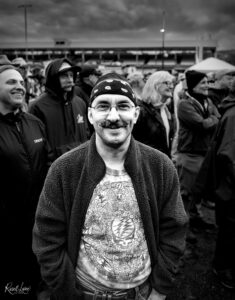 Man with a mustache wearing a bandana and glasses in the foreground, smiling as a crowd stands behind him at an outdoor event (black and white).