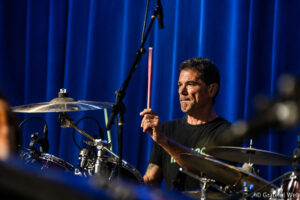 Drummer on stage mid-performance, raised drumstick over blue backstage curtains and drum kit visible to the left side