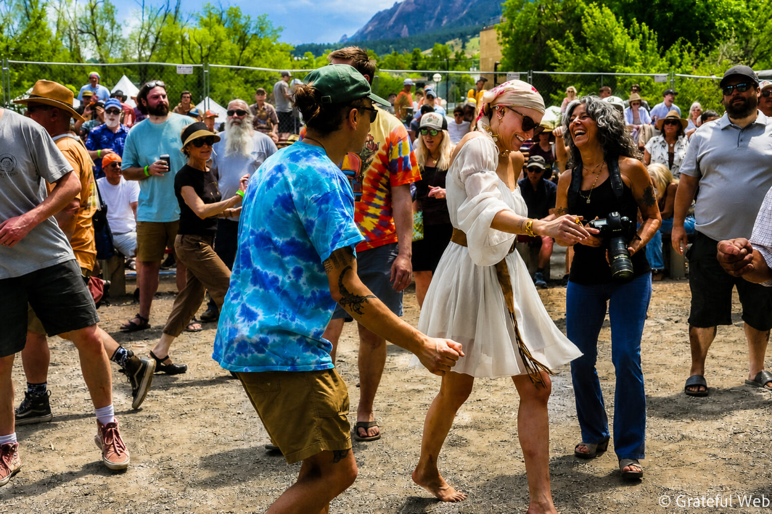 Crowd of festival-goers dancing outdoors, colorful clothing and smiles with mountains in the background.