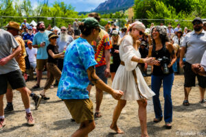 Crowd of festival-goers dancing outdoors, colorful clothing and smiles with mountains in the background.