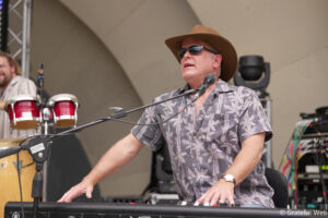 Man wearing a brown wide-brim hat and sunglasses plays a keyboard on stage, with a microphone in front.