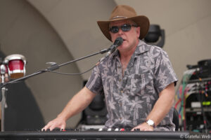Man wearing a brown cowboy hat and sunglasses sings into a microphone while playing a keyboard on stage.